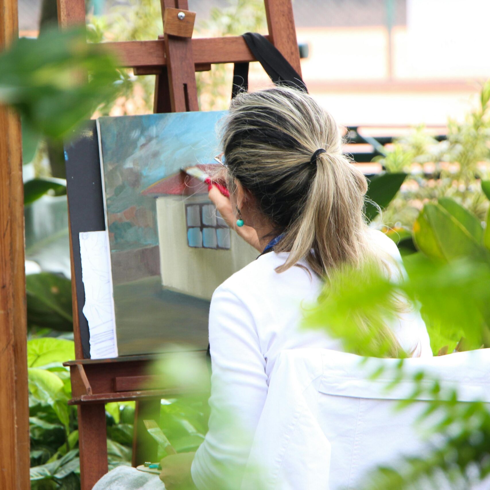 Home A woman enjoys painting in a lush outdoor garden, surrounded by vibrant artwork.